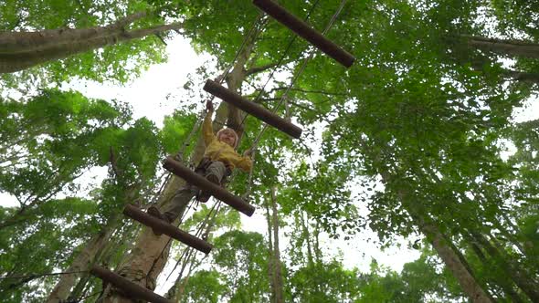 Slowmotion Shot of a Little Boy in a Safety Harness Climbs on a Route in Treetops in a Forest