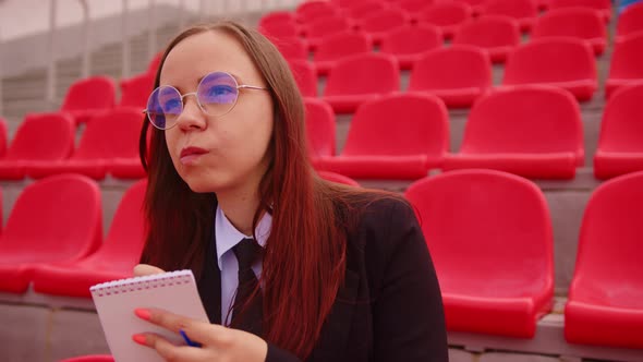 Young Woman in Glasses with Notepad Pen Sitting on Stadium Bleachers Alone