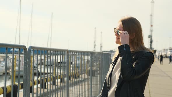 Beautiful Blonde in Black Leather Jacket in Port Against Background of Boats and Ships Raises Her