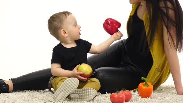 Mom and Son Are Sitting on the Floor Holding Vegetables. White Background