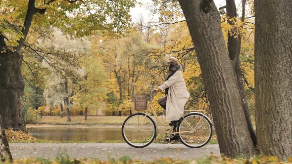 Young Stylish Woman in a Beige Raincoat Rides a Bicycle Through an Autumn Park in Slow Motion