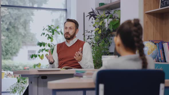 Male Teacher Sitting at the Desk and Actively Explaining a Topic