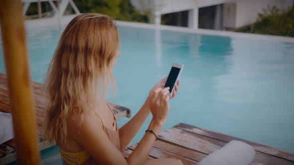 A Blonde Woman Uses Her Mobile Phone While Lounging in the Sun By the Pool on Vacation