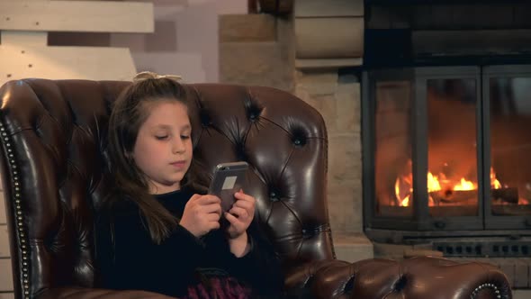 Little Girl Spend Her Leisure Time with Smartphone Near the Fireplace