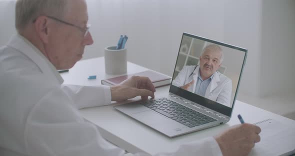 Two Colleagues Doctors Are Communicating By Video Call on Laptop, Professor Is Writing Notes in His