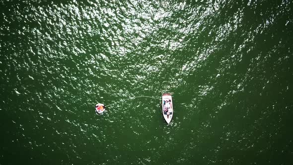 Drone shot zooming in on a boat staying still on a green lake. The boat has a rope leading to a tube