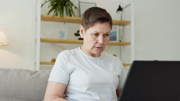 Smiling Elderly Mature Woman Resting on Sofa Using Laptop Alone at Home