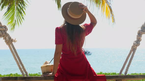 Tourist Woman in Hat and Red Dress Sitting on Wooden Swing with Young Coconut and Enjoy Sea and Palm