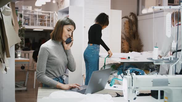 Woman Clarifying Order Details By Smartphone Working at Sewing Workshop Mixed Race Seamstresses