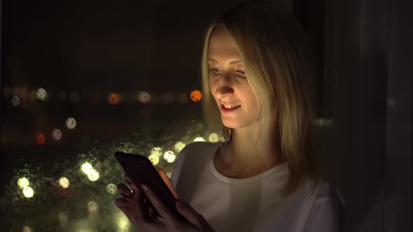 young Caucasian lady stands at the window at night, reading the news on phone.