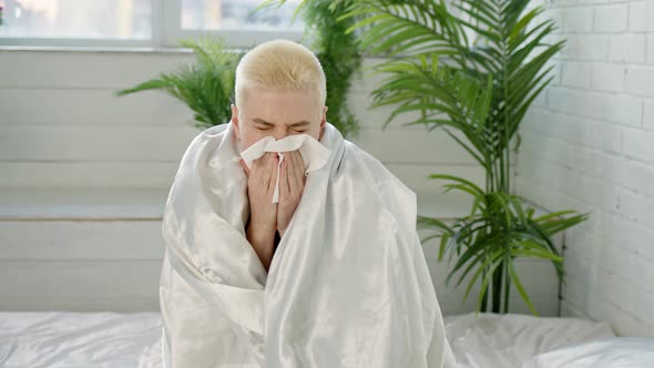 Upset Woman with Short White Hair Wrapped in White Blanket at Home Blows Her Runny Nose Into