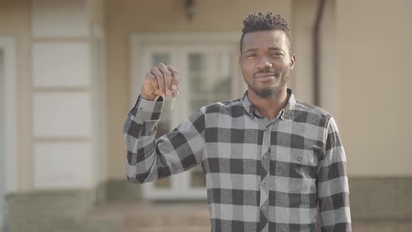 Handsome African American Man in Checkered Shirt Standing in Front of a Big House Holding Keys