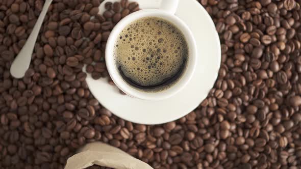 White Cup, Saucer, Spoon On Background Of Coffee Beans. Cup Fresh Espresso With Froth Coffee In Bag