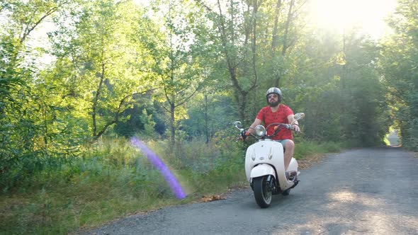 Bearded Hipster Rides Past on a Vintage Scooter in the Forest. Handsome Man Rides on a Country Road