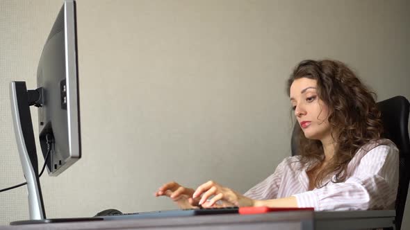 Tired Young Woman with Curly Hair and White Shirt is Working at the Office Using Her Computer