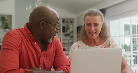 Happy senior diverse couple wearing shirts and using laptop in living room