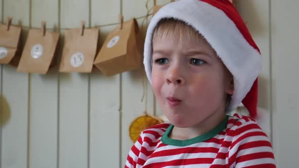 Boy in Pajamas Santa Cap Eating Sweet Candy on Paper Advent Calendar Background