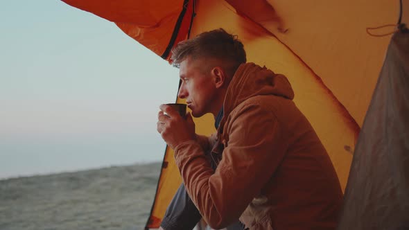Adult Man Hiker Sitting at Tent Entry and Holding Mug with Coffee in Hands