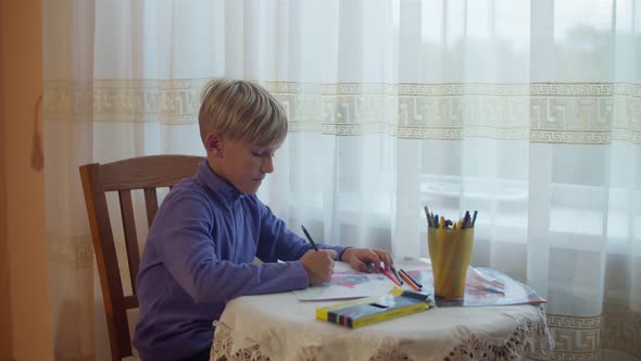 A schoolboy boy draws at a table by the window. The boy sits in the room and draws