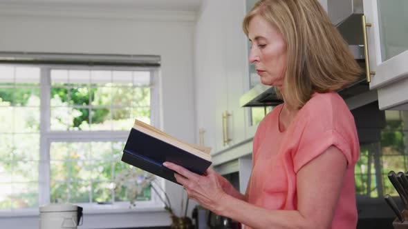 Caucasian senior woman standing in kitchen at home reading book