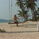 A Blonde with Long Hair in a Swimsuit Sways on a Swing Suspended From a Palm Tree Back View - VideoHive Item for Sale