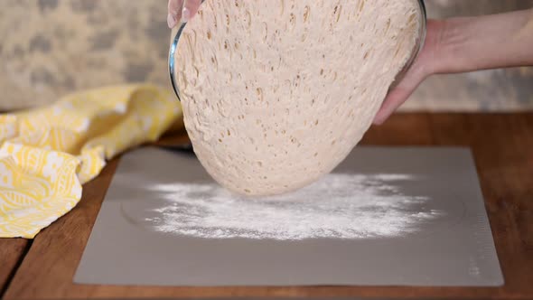 A baker's hands pouring bread dough from a bowl onto a kitchen table covered in flour