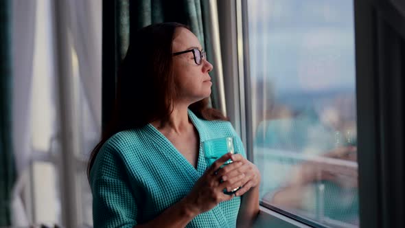 Middleaged Woman at Home in Morning Looking at Window Holding Glass of Water