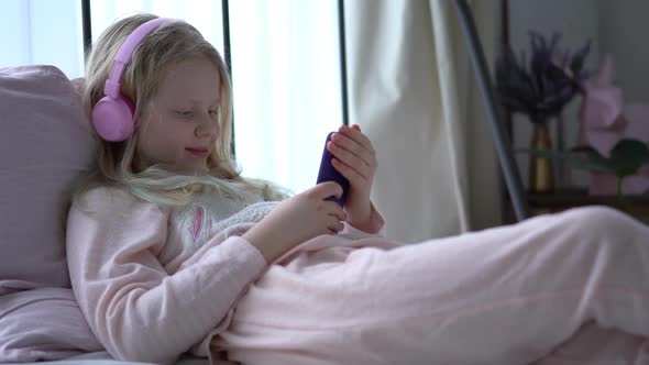 Modern Life of Generation Z. Teenage Girl in Pajamas and Headphones in the Room on the Bed Listens