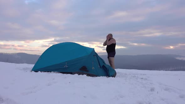 Hiker in shorts drink tea from thermos in front of tent, in winter mountains