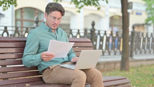 Man Reading Documents while working on Laptop