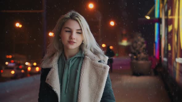 Girl Walks and Smiles Winter Night During Snowfall Near Glowing Shopping Center