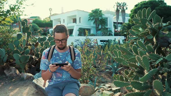 Bearded Man Is Resting in Park in Summer Day, Sitting on Bench and Playing Phone