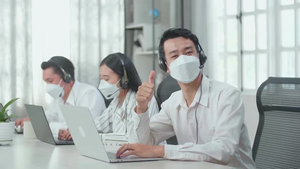 A Man Of Three Asian Call Centre Agents Wearing Mask, Looking To Camera And Thumbs Up