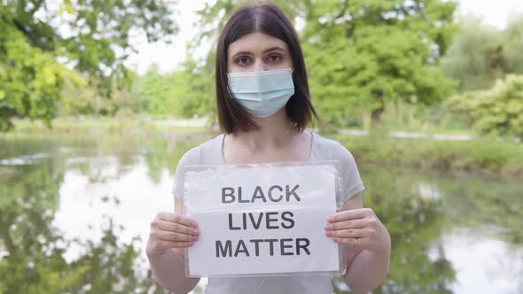 A Young Caucasian Woman in a Face Mask Shows a Black Lives Matter Sign to the Camera in a Park