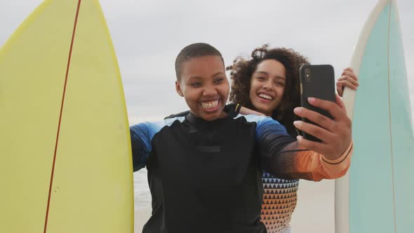 Happy african american female friends on the beach holding surfboards taking selfie