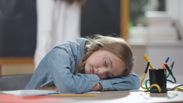 Tired Caucasian Schoolgirl Sleeping on Desk in Classroom with Blurred Classmate Jumping at