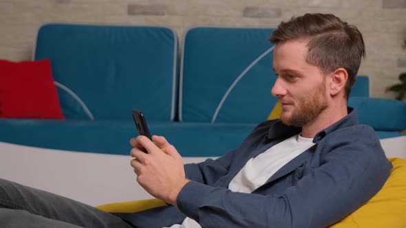 Young Man Sits on Bean Bag and Playing Games on Phone, at Home