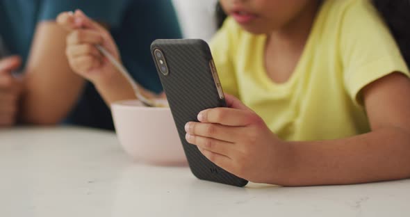 Happy biracial father and daughter eating breakfast, using smartphone