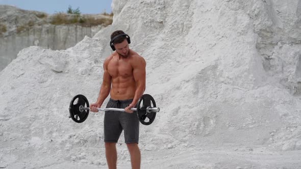 Bodybuilder lifting barbell and looking on his muscular body outside.