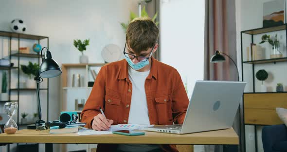 Young Male Freelancer or Worker in Facial Medical Mask Working on Laptop at Home, Covid-19