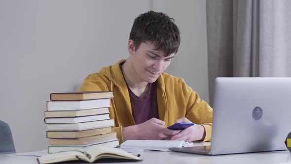 Smiling Brunette Caucasian Boy Sitting at the Table with Books and Laptop and Using Smartphone