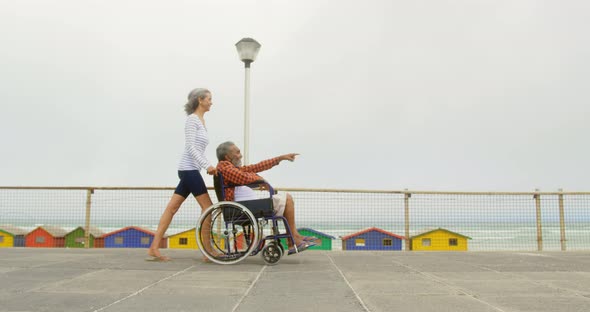 Side view of active senior African American woman pushing disabled man in wheelchair on promenade 4k