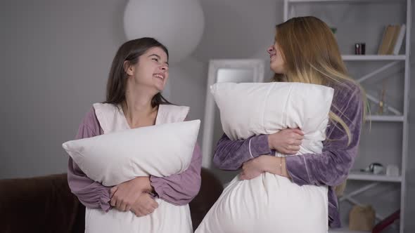 Medium Shot Portrait of Two Happy Beautiful Caucasian Women in Pajamas Standing with Soft Pillows