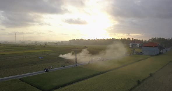 Aerial drone view of farming fields and a road with burning smoke at sunset.