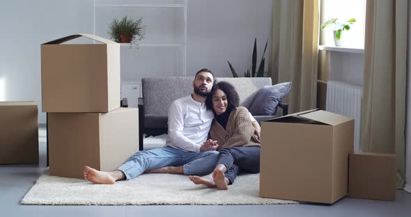 Tired Multietnical Couple Hugging Kissing Sit on Floor in Living Room Surrounded By Big Carton Boxes