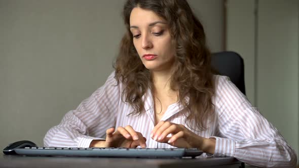Young Female Office Manager in White Shirt and Curly Hair is Sitting at the Table and Typing Using