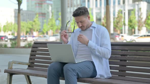 Man with Headache Using Laptop While Sitting Outdoor on Bench