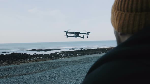 Close-up Man in Hat Is Controlling Drone with Remote, Flying Very Close at Gorgeous Calm Sea Beach