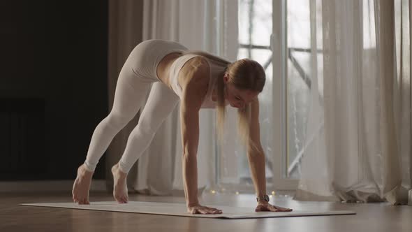 A Young Woman in White Sportswear is Stretching with a Large Hall with Large Windows in a Slowmotion