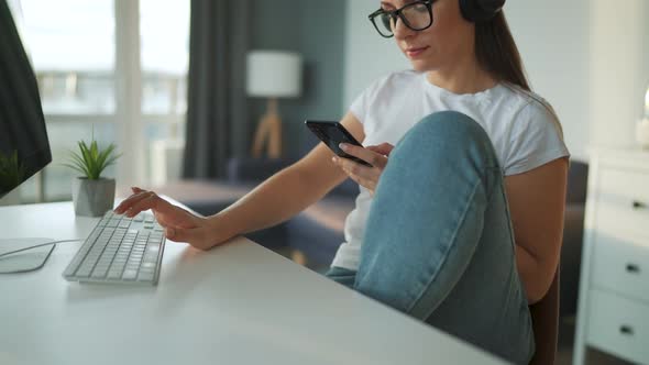 Casually Dressed Woman with Headphones Working with a Computer and Smartphone at Home in a Cozy